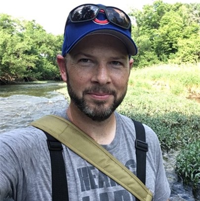 Man outdoors near a stream wearing a cap and shoulder strap.